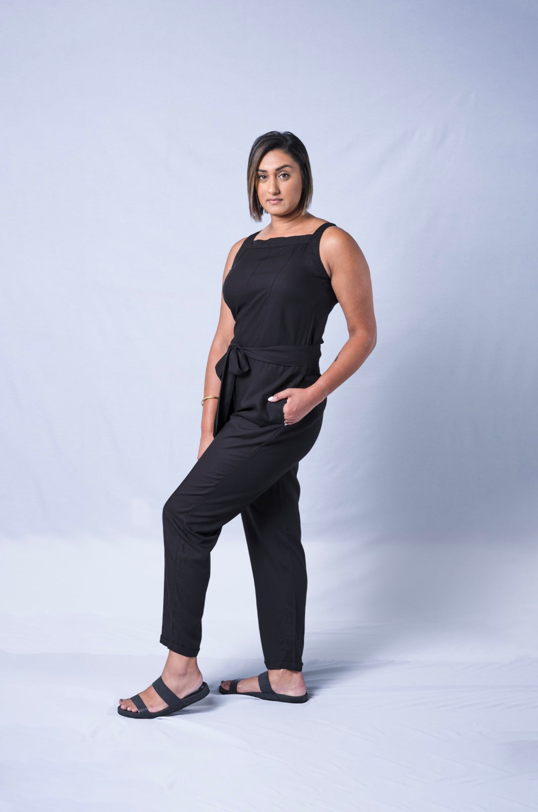 All In One Jumpsuit Model Standing showing side of the jumpsuit. Waistband tied to front and hand in pocket